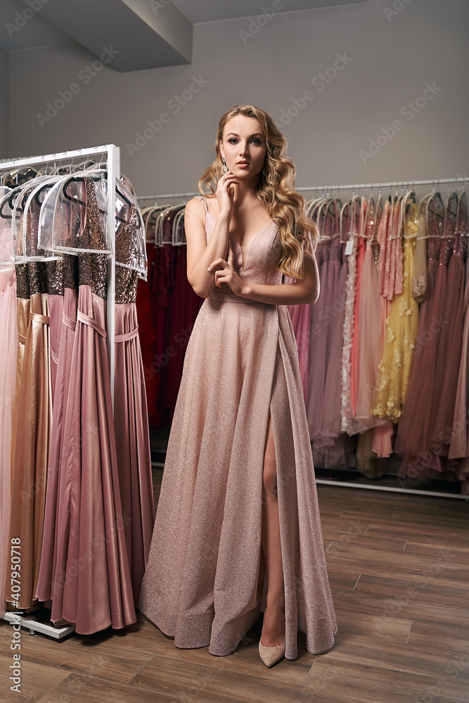 Young beautiful blonde girl wearing a full-length pale pink glitter chiffon draped prom ball gown. Model selecting an outfit for occasion in dress hire service with many options on background. Young beautiful blonde girl wearing a full-length pale pink glitter chiffon draped prom ball gown. Model selecting an outfit for occasion in dress hire service with many options on background.