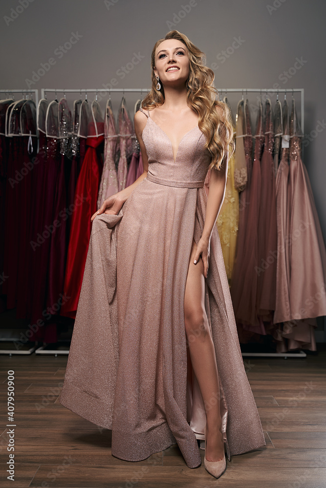Young beautiful blonde girl wearing a full-length pale pink glitter chiffon draped prom ball gown. Model selecting an outfit for occasion in dress hire service with many options on background.