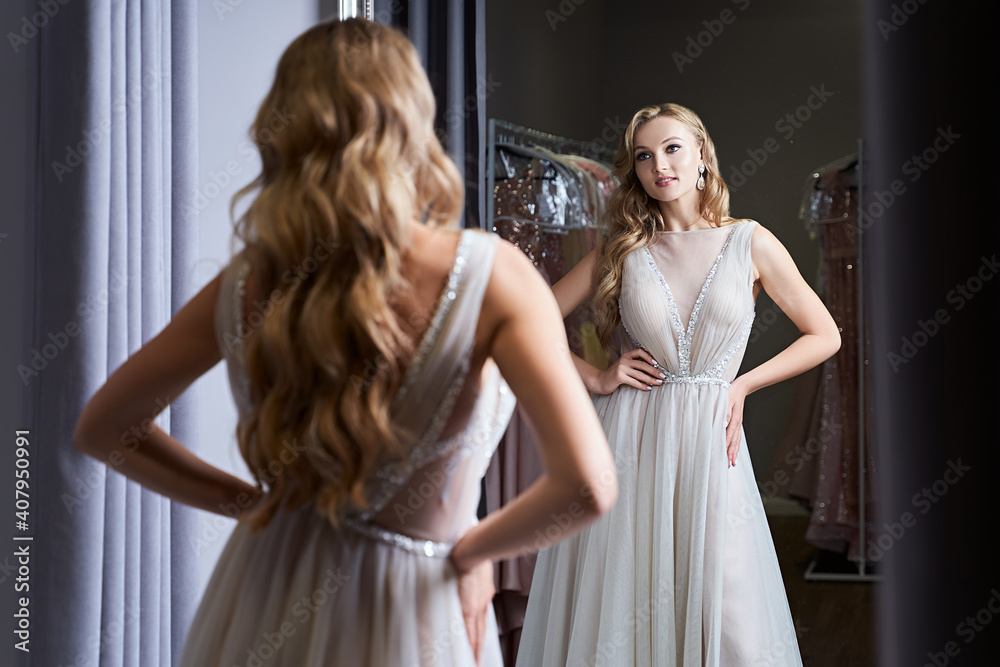 Young beautiful blonde girl wearing a full-length silver white chiffon prom ball gown decorated with sparkles and sequins. Model in front of mirror in a fitting room at dress hire service.