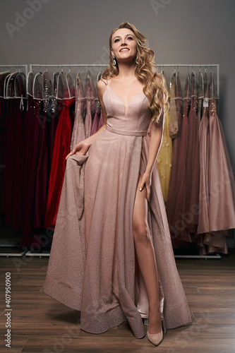 Obraz na płótnie Young beautiful blonde girl wearing a full-length pale pink glitter chiffon draped prom ball gown. Model selecting an outfit for occasion in dress hire service with many options on background. w salonie