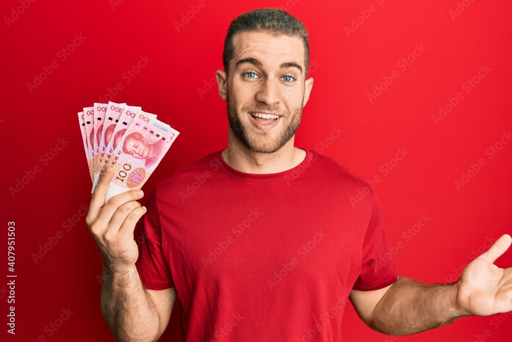 Young caucasian man holding yuan chinese banknotes celebrating achievement with happy smile and winner expression with raised hand Young caucasian man holding yuan chinese banknotes celebrating achievement with happy smile and winner expression with raised hand