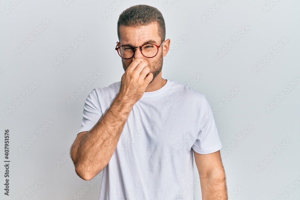 Young caucasian man wearing casual clothes and glasses smelling something stinky and disgusting, intolerable smell, holding breath with fingers on nose. bad smell Young caucasian man wearing casual clothes and glasses smelling something stinky and disgusting, intolerable smell, holding breath with fingers on nose. bad smell