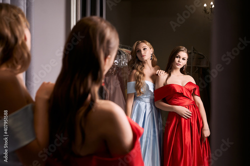 Two young beautiful girls wearing off-the-shoulder full-length sky blue and crimson red satin slit prom ball gowns. Models looking in mirror. Fitting room in dress hire service.