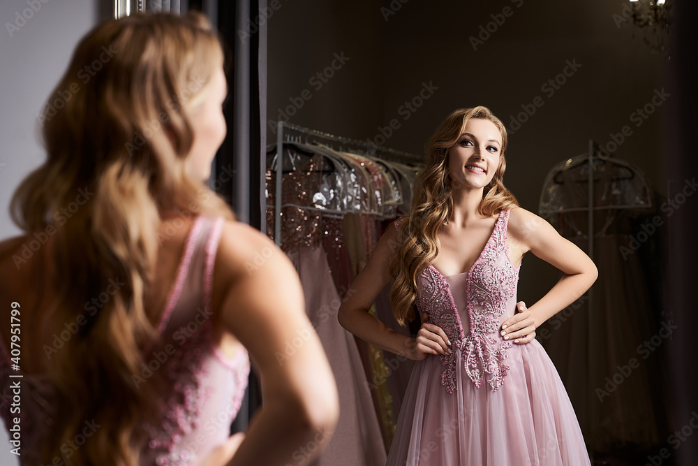Young beautiful blonde girl wearing a full-length pink violet or purple chiffon prom ball gown decorated with sparkles and sequins. Model in front of mirror in a fitting room at dress hire service.