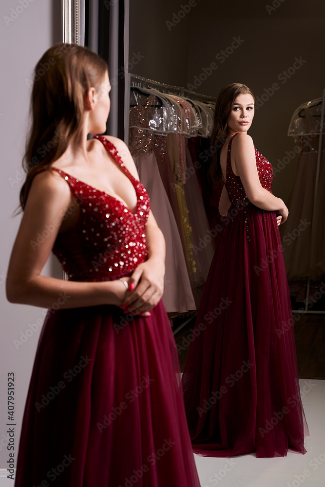 Young beautiful brunette girl wearing a full-length dark crimson red chiffon prom ball gown decorated with sparkles and sequins. Model in front of mirror in a fitting room at dress hire service. Young beautiful brunette girl wearing a full-length dark crimson red chiffon prom ball gown decorated with sparkles and sequins. Model in front of mirror in a fitting room at dress hire service.