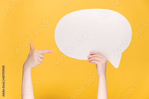 Close up of unrecognizable young female holding and pointing at blank white paper speech bubble, isolated on yellow background with copy space for advertisement. Body language concept
