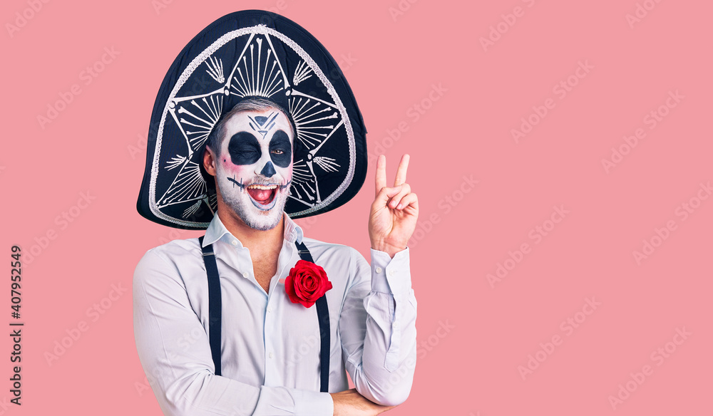 Man wearing day of the dead costume over background smiling with happy face winking at the camera doing victory sign with fingers. number two.