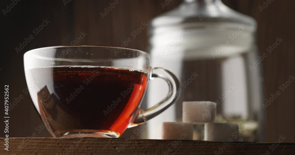 Hot tea in clear glass Cup closeup, slow motion. Next to it are pieces of refined sugar. A can of hand-picked herbal tea stands against a wooden background. Tea ceremony. There is steam from hot tea. Hot tea in clear glass Cup closeup, slow motion. Next to it are pieces of refined sugar. A can of hand-picked herbal tea stands against a wooden background. Tea ceremony. There is steam from hot tea.