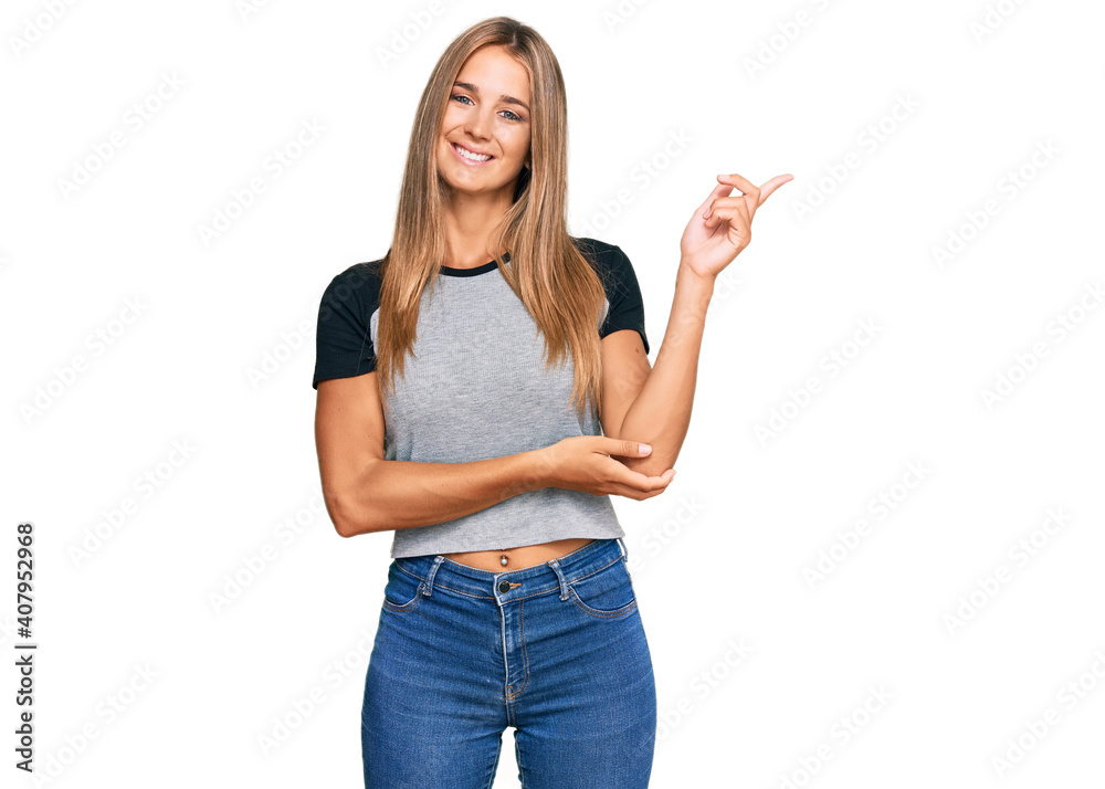 Young blonde woman wearing casual clothes with a big smile on face, pointing with hand and finger to the side looking at the camera.