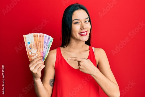 Young hispanic girl holding philippine peso banknotes smiling happy pointing with hand and finger