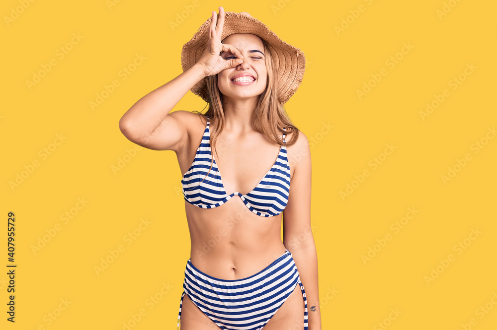 Young beautiful blonde woman wearing bikini and hat doing ok gesture with hand smiling, eye looking through fingers with happy face. Young beautiful blonde woman wearing bikini and hat doing ok gesture with hand smiling, eye looking through fingers with happy face.