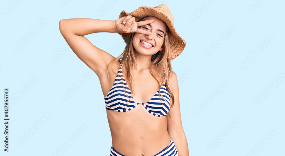 Young beautiful blonde woman wearing bikini and hat doing peace symbol with fingers over face, smiling cheerful showing victory Young beautiful blonde woman wearing bikini and hat doing peace symbol with fingers over face, smiling cheerful showing victory