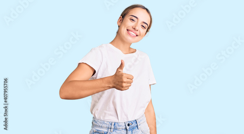 Young beautiful blonde woman wearing casual white tshirt doing happy thumbs up gesture with hand. approving expression looking at the camera showing success. photo