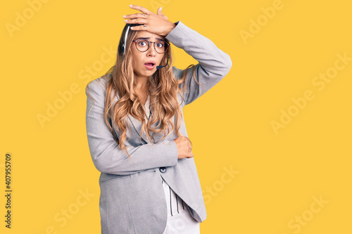Young beautiful blonde woman wearing call center agent headset surprised with hand on head for mistake, remember error. forgot, bad memory concept.