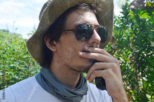 Young farmer smoking in nature. Relaxing and smoking outside. Casual farmer taking a break. Attractive man with facial hair smoking outdoors. Young male hiker resting.