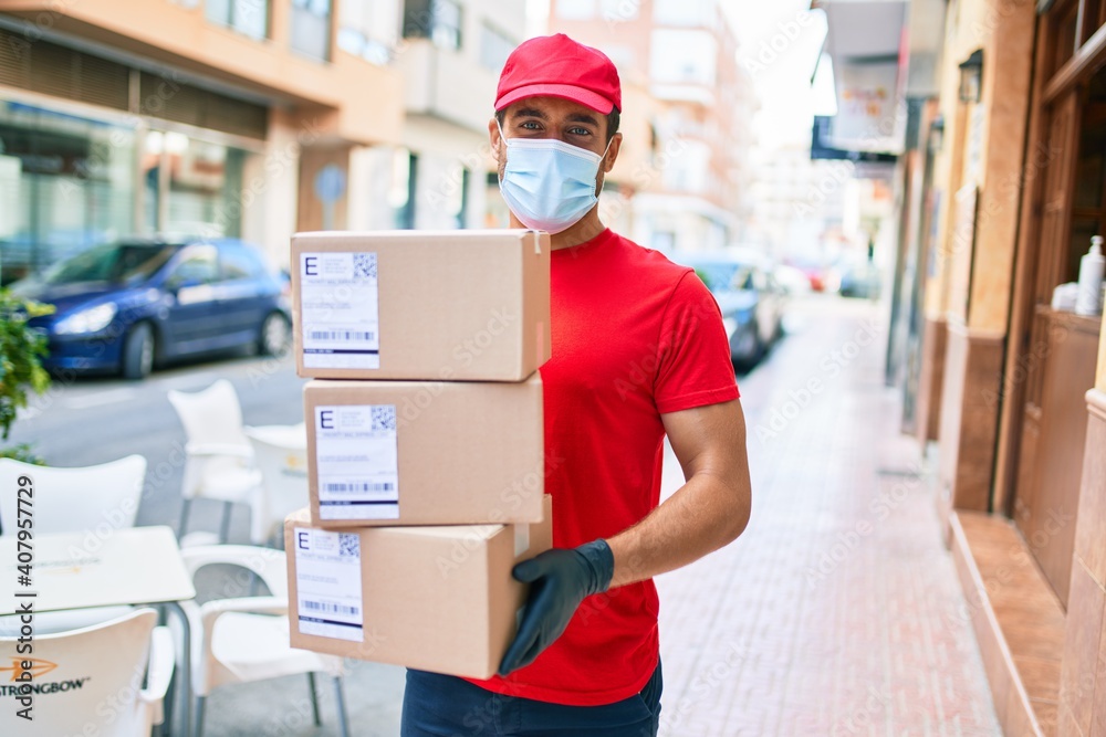 Young delivery man wearing uniform and coronavirus protection medical mask holding cardboard packages at town street. Young delivery man wearing uniform and coronavirus protection medical mask holding cardboard packages at town street.