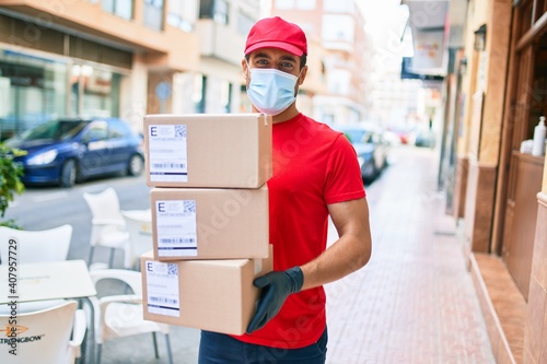 Young delivery man wearing uniform and coronavirus protection medical mask holding cardboard packages at town street.