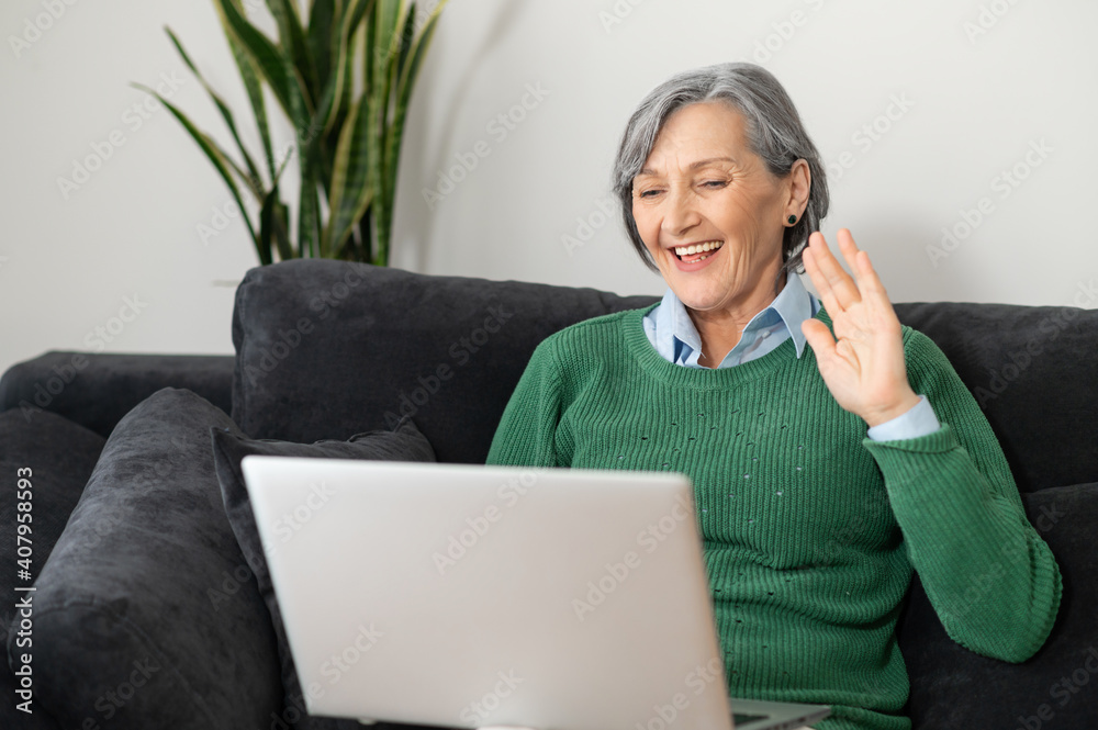 A smiling grandmother wearing a green jumper is sitting in the living room, relaxing and talking to her friend or family, waving at the laptop screen, staying connected during pandemic A smiling grandmother wearing a green jumper is sitting in the living room, relaxing and talking to her friend or family, waving at the laptop screen, staying connected during pandemic