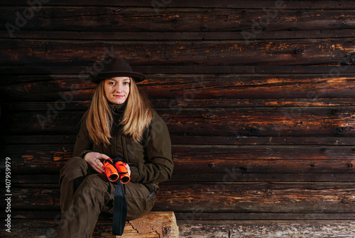 A young beautiful hunter woman sits with a telescope in her hands. The background is an old wooden log cabin.