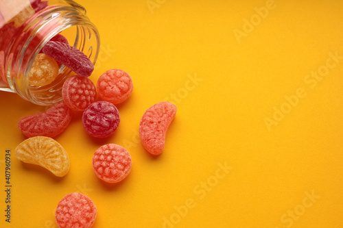 Sweet fruit hard candies pouring out of the glass mason jar on a colored yellow background. Overhead view of colorful caramel assortment.