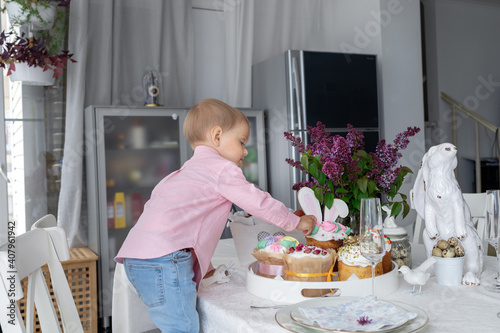  little cute white boy is standing at the Easter table and eating Easter cakes, lilac, Easter bunny, Easter decorations, painted Easter eggs. High quality photo