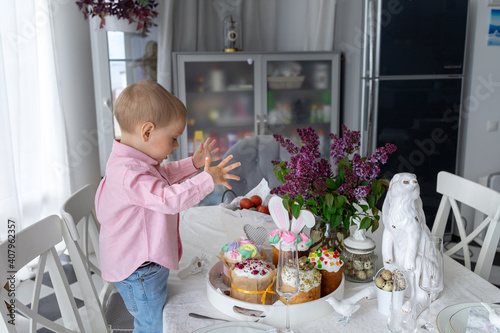  little cute white boy is standing at the Easter table and eating Easter cakes, lilac, Easter bunny, Easter decorations, painted Easter eggs. High quality photo