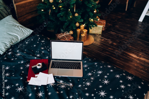 Modern laptop on comfortable bed against shiny Christmas tree