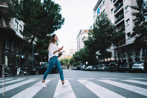 Smiling caucasian female in casual wear walking on zebra in city holding mobile phone with navigation application, positive woman 20s millennial crossing road using smartphone unattentive in downtown photo