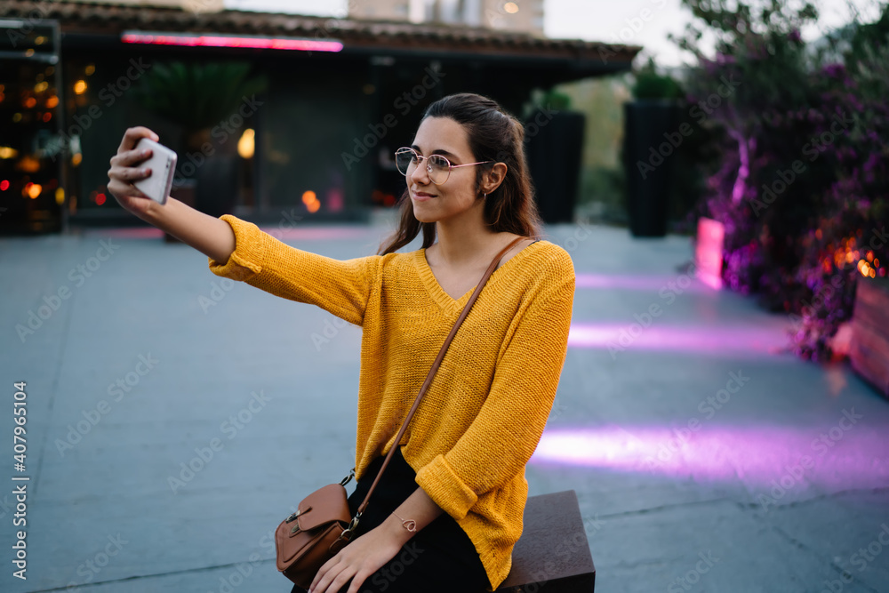 Fototapeta premium Positive trendy dressed caucasian woman in eyewear holding mobile phone posing for selfie on free time recreating, attractive female with smartphone shooting video for share in social networks via 4G