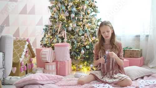 Adorable little girl sitting near the tree and making paper snow-flakes. Room decorated. photo