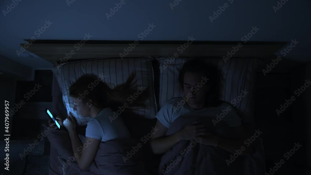 Young couple in bed at night, man is asleep, woman is looking at smartphone, female insomnia concept, top view. Young couple in bed at night, man is asleep, woman is looking at smartphone, female insomnia concept, top view.