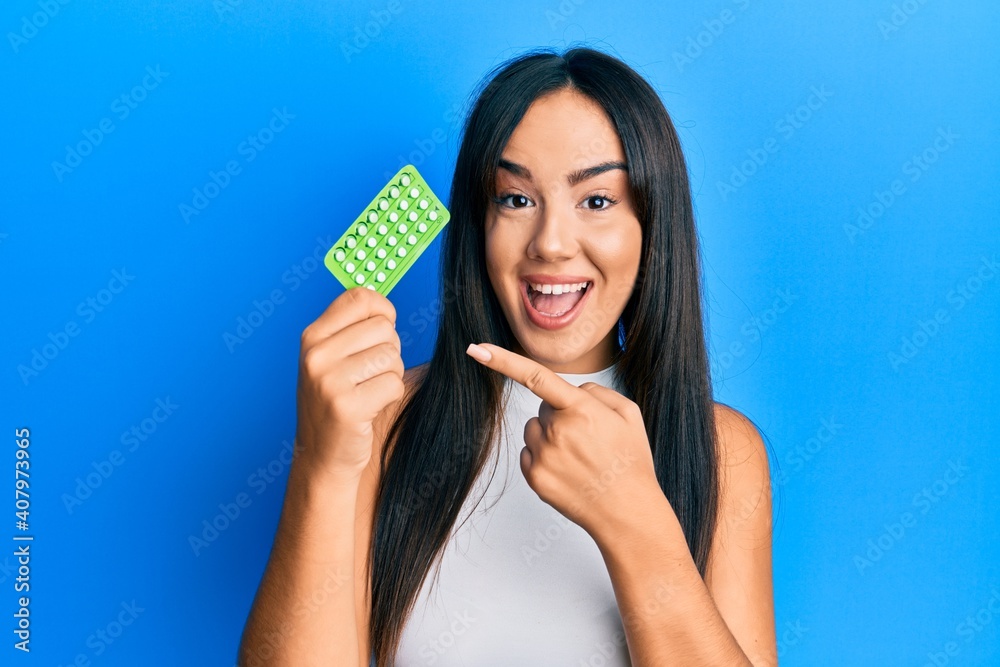 Young beautiful hispanic girl holding birth control pills smiling happy pointing with hand and finger Young beautiful hispanic girl holding birth control pills smiling happy pointing with hand and finger