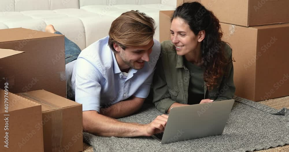 Smiling millennial family couple lying on floor between huge carton boxes, discussing interior decoration purchases searching stuff in online store using computer, renovation relocation concept. Smiling millennial family couple lying on floor between huge carton boxes, discussing interior decoration purchases searching stuff in online store using computer, renovation relocation concept.