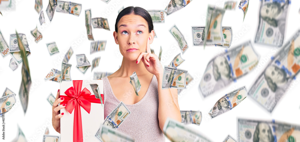Beautiful brunette young woman holding gift serious face thinking about question with hand on chin, thoughtful about confusing idea Beautiful brunette young woman holding gift serious face thinking about question with hand on chin, thoughtful about confusing idea