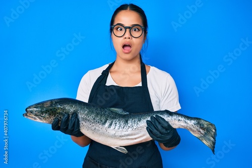 Young beautiful asian girl fishmonger selling fresh raw salmon afraid and shocked with surprise and amazed expression, fear and excited face.