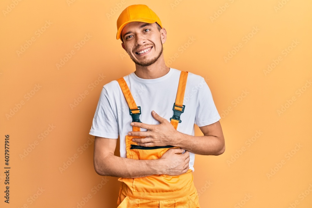Hispanic young man wearing handyman uniform smiling and laughing hard out loud because funny crazy joke with hands on body.