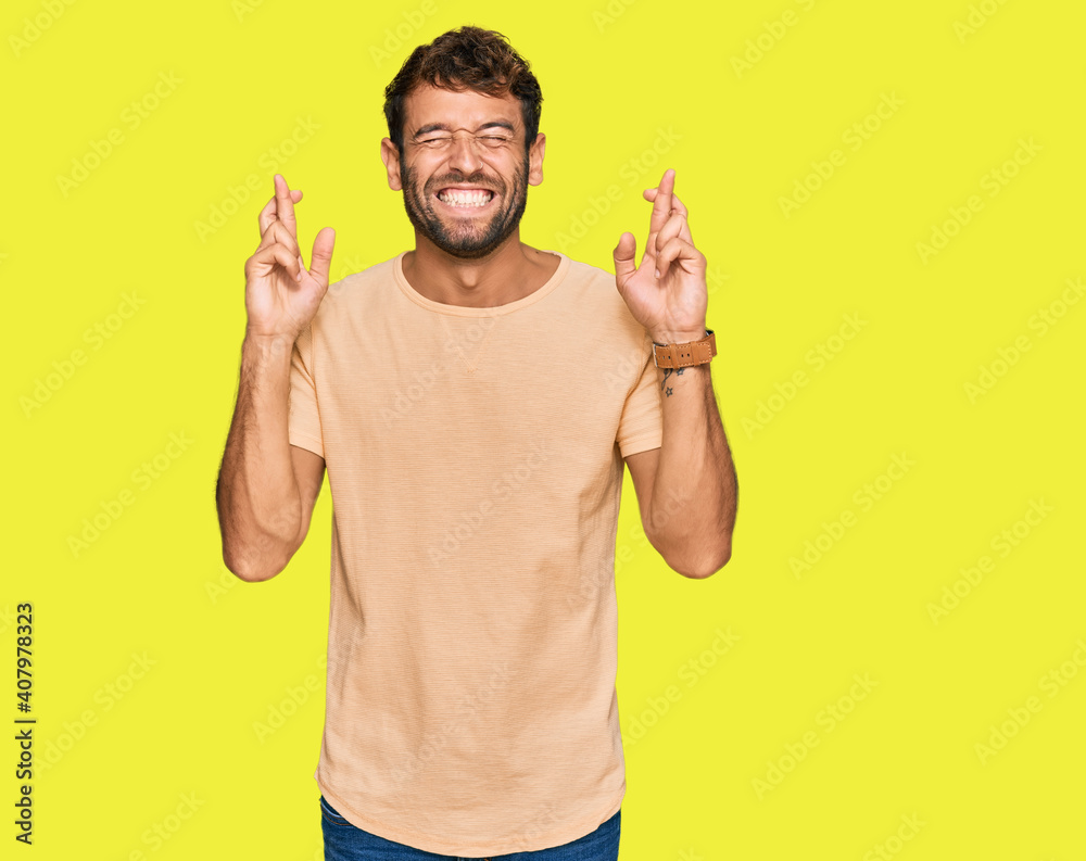 Handsome young man with beard wearing casual tshirt gesturing finger crossed smiling with hope and eyes closed. luck and superstitious concept. Handsome young man with beard wearing casual tshirt gesturing finger crossed smiling with hope and eyes closed. luck and superstitious concept.