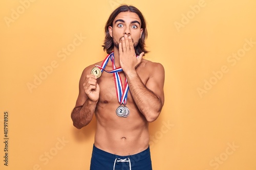 Young handsome man wearing swimwear and medals covering mouth with hand, shocked and afraid for mistake. surprised expression