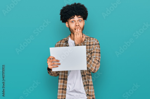 Young african american man with afro hair holding blank empty banner covering mouth with hand, shocked and afraid for mistake. surprised expression