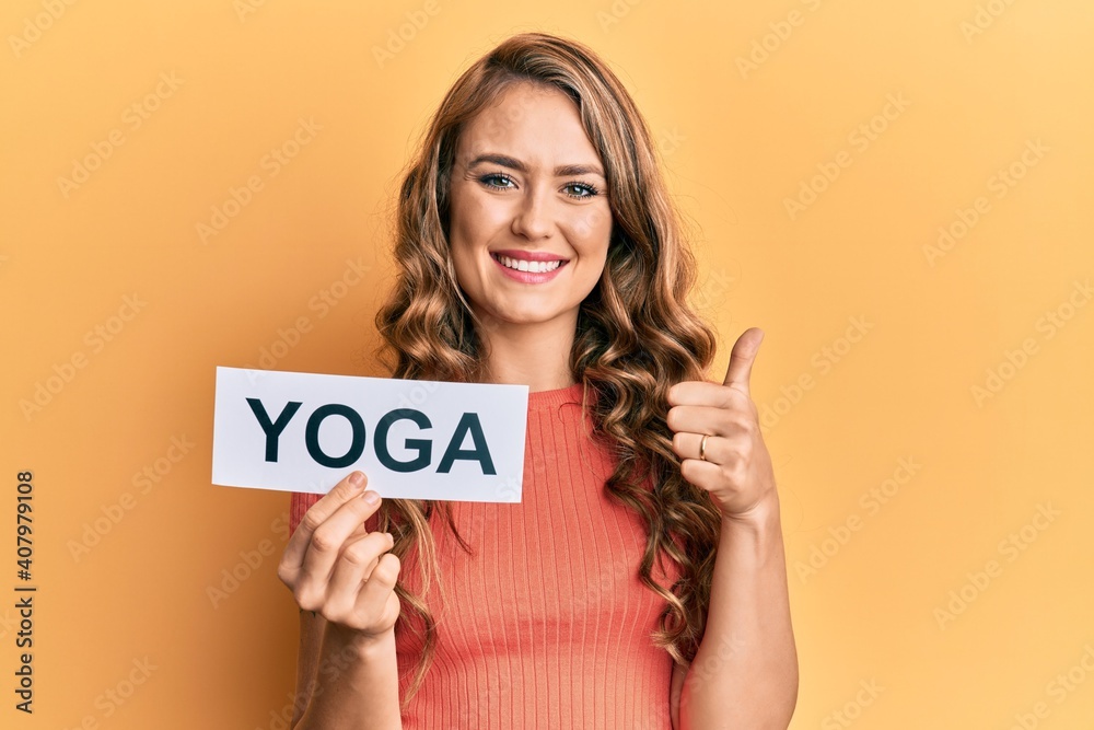 Young blonde girl holding yoga message paper smiling happy and positive, thumb up doing excellent and approval sign Young blonde girl holding yoga message paper smiling happy and positive, thumb up doing excellent and approval sign