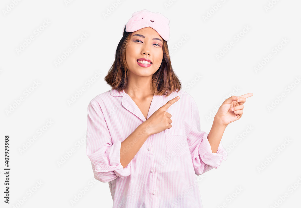 Young beautiful chinese girl wearing sleep mask and pajama smiling and looking at the camera pointing with two hands and fingers to the side. Young beautiful chinese girl wearing sleep mask and pajama smiling and looking at the camera pointing with two hands and fingers to the side.