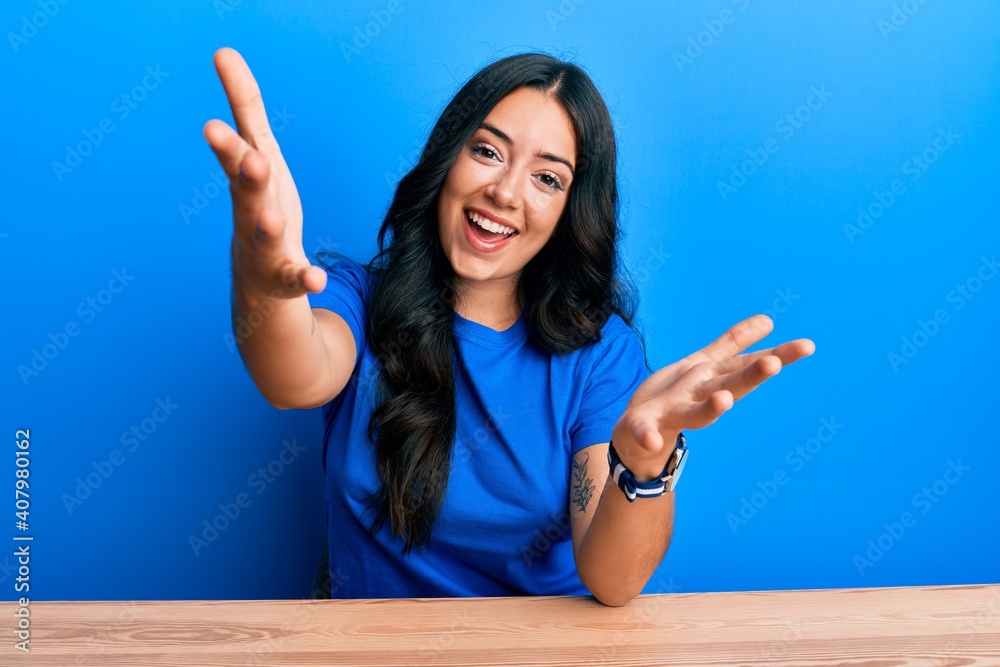Beautiful brunette young woman wearing casual clothes sitting on the table looking at the camera smiling with open arms for hug. cheerful expression embracing happiness. Beautiful brunette young woman wearing casual clothes sitting on the table looking at the camera smiling with open arms for hug. cheerful expression embracing happiness.