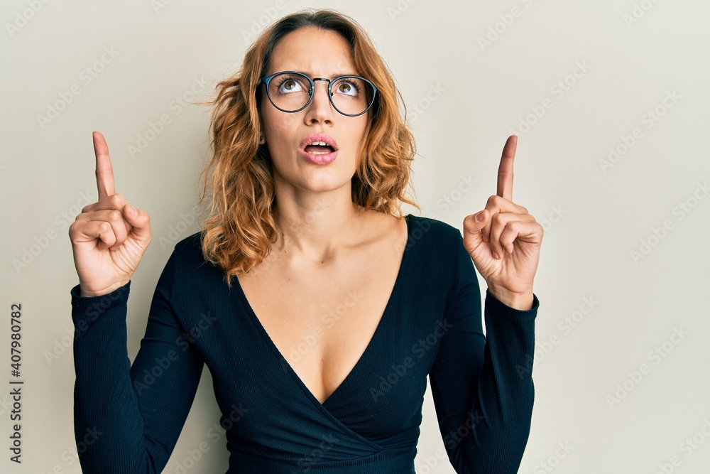 Young caucasian woman wearing business shirt and glasses amazed and surprised looking up and pointing with fingers and raised arms. Young caucasian woman wearing business shirt and glasses amazed and surprised looking up and pointing with fingers and raised arms.