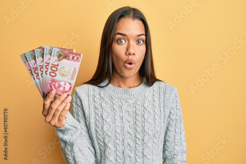 Beautiful hispanic woman holding 100 new zealand dollars banknote scared and amazed with open mouth for surprise, disbelief face