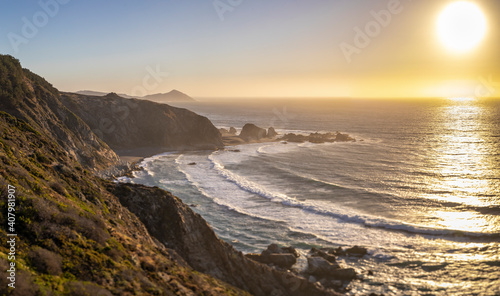 An awe sunset in an idyllic beach landscape with the sunset over the water and the sunlight generating a moody atmosphere. An amazing wil scenery with the cliffs and the sea waves splashing water