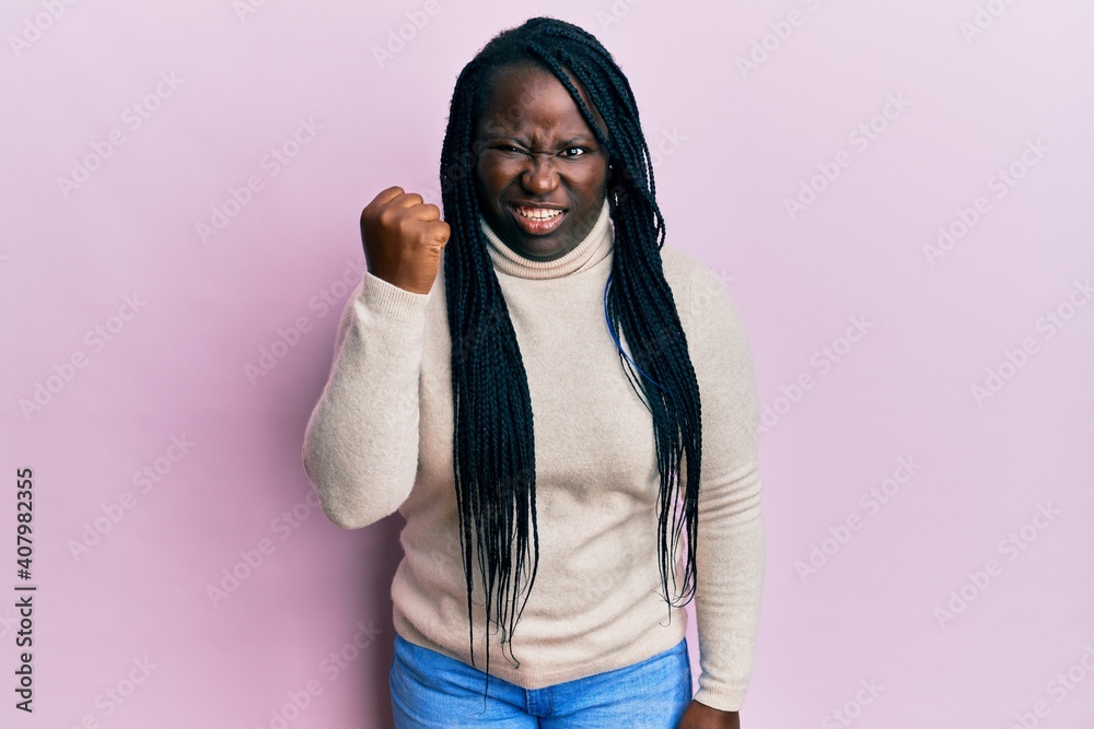 Young black woman with braids wearing casual winter sweater angry and mad raising fist frustrated and furious while shouting with anger. rage and aggressive concept. Young black woman with braids wearing casual winter sweater angry and mad raising fist frustrated and furious while shouting with anger. rage and aggressive concept.