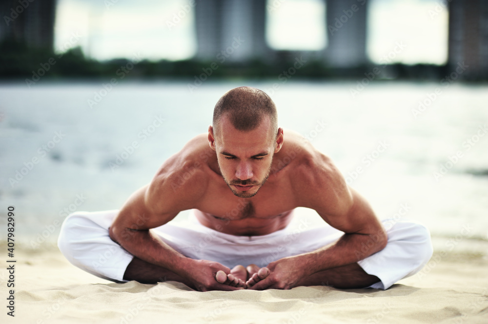 Obraz premium Young caucasian male practicing yoga in white pants on a sandy shore.