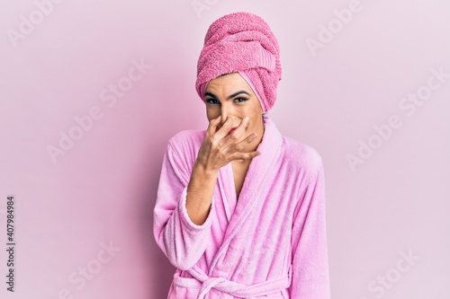 Young man wearing woman make up wearing shower towel on head and bathrobe smelling something stinky and disgusting, intolerable smell, holding breath with fingers on nose. bad smell © Krakenimages.com