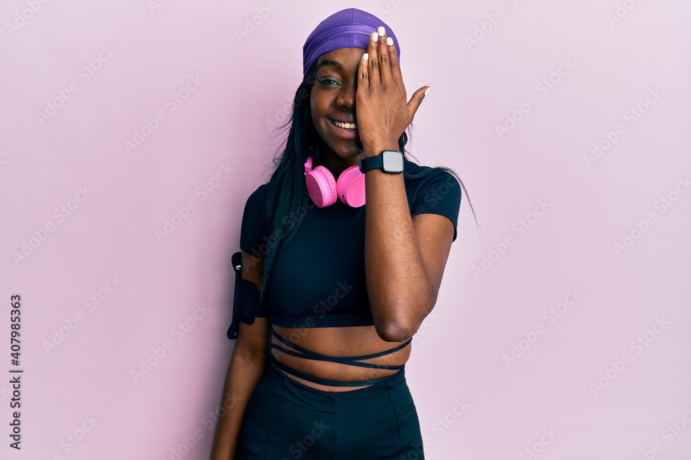 Young african american woman wearing gym clothes and using headphones covering one eye with hand, confident smile on face and surprise emotion. Young african american woman wearing gym clothes and using headphones covering one eye with hand, confident smile on face and surprise emotion.