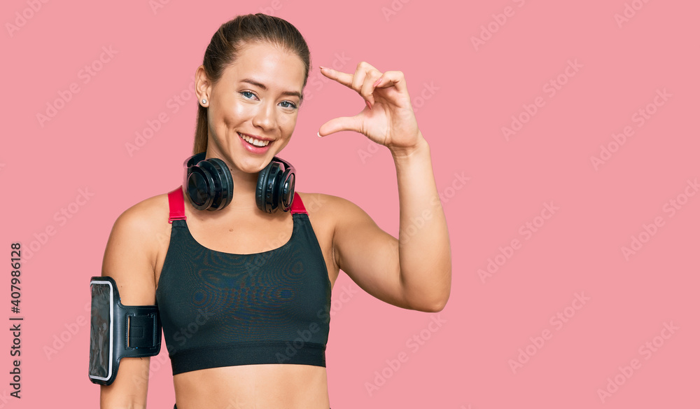 Beautiful blonde woman wearing gym clothes and using headphones smiling and confident gesturing with hand doing small size sign with fingers looking and the camera. measure concept. Beautiful blonde woman wearing gym clothes and using headphones smiling and confident gesturing with hand doing small size sign with fingers looking and the camera. measure concept.